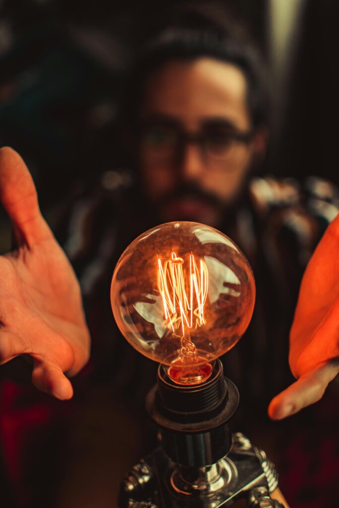 A man with glasses focuses on a glowing light bulb in a dim indoor setting.